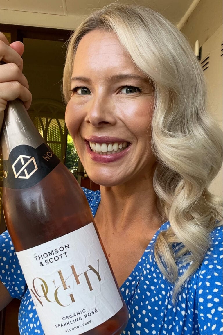A smiling woman with long blond hair wearing a blue polka dot dress holds up a bottle of Thomson & Scott Noughty non-alcoholic sparkling rosé wine. She stands indoors in front of an open door.
