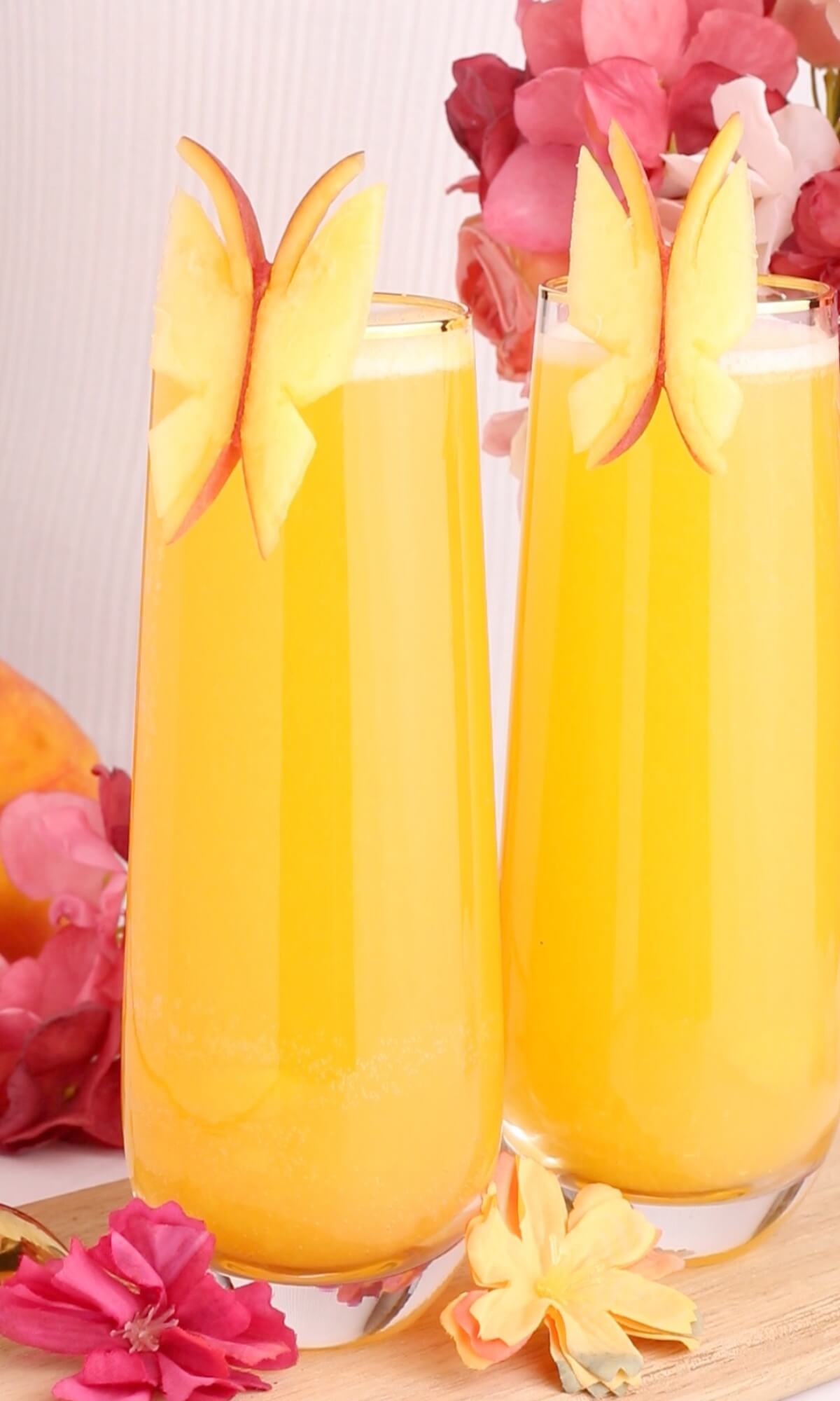 Two tall glasses of bright yellow juice are garnished with butterfly-shaped apple slices. Pink and peach flowers surround the glasses, creating a colorful and fresh presentation.