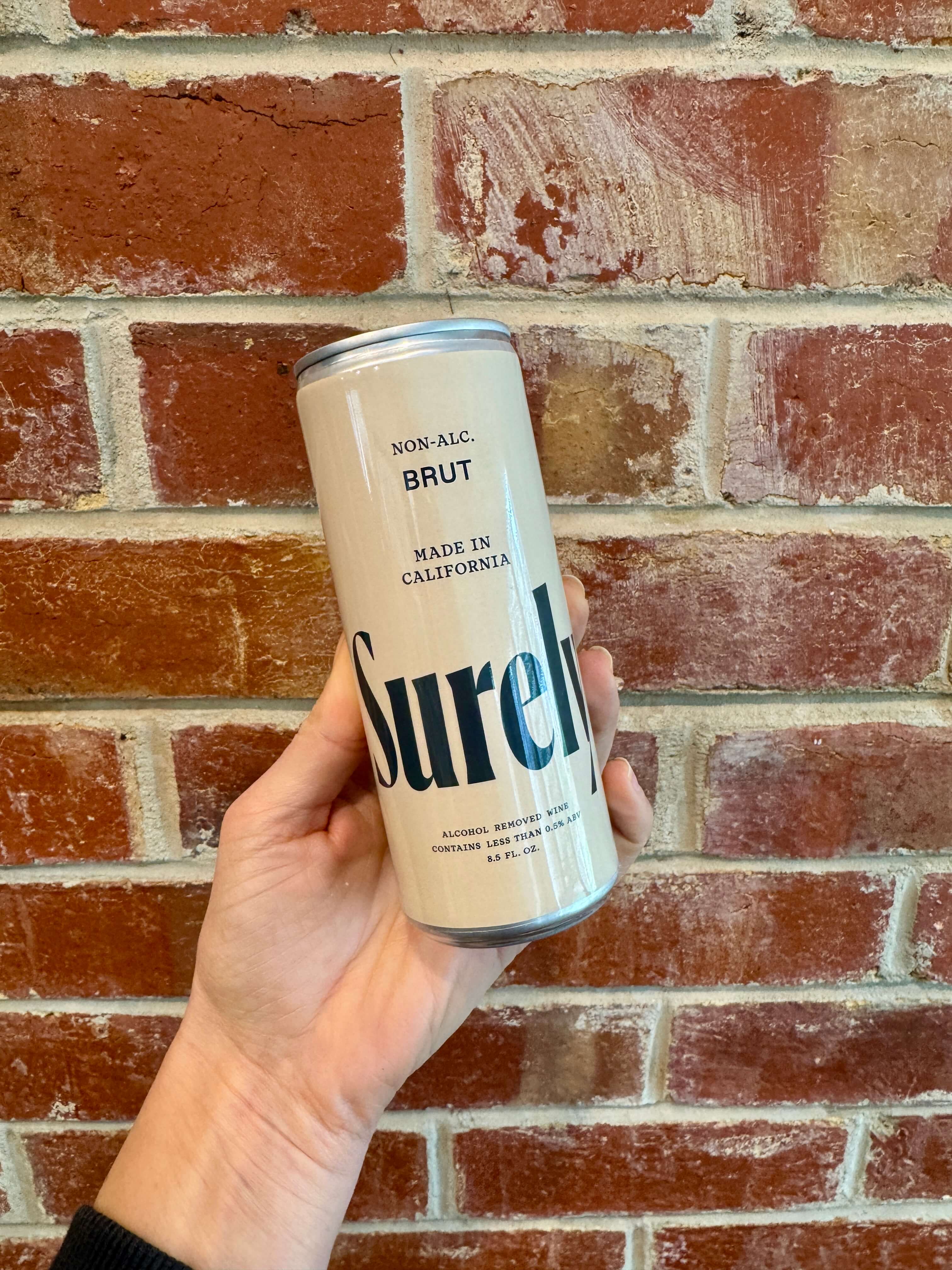 A hand holds a beige can labeled Surely Non-Alc. Brut, Made in California in front of a red brick wall.