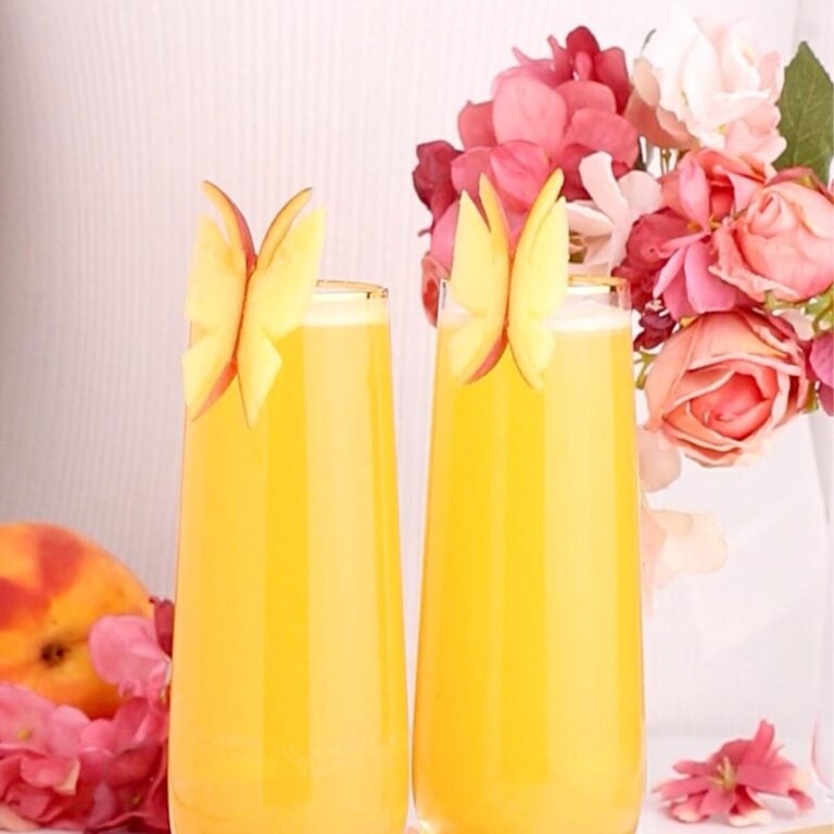 Two tall glasses of yellow-orange juice are garnished with butterfly-shaped apple slices. Pink flowers and a peach are in the background, creating a fresh and colorful scene.
