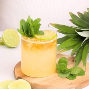 A refreshing yellow cocktail garnished with a lime wheel and mint sprig, served in a glass of ice on a wooden board with fresh lime slices, mint leaves, and a pineapple nearby.