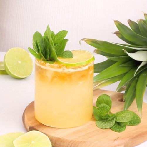 A refreshing yellow cocktail garnished with a lime wheel and mint sprig, served in a glass of ice on a wooden board with fresh lime slices, mint leaves, and a pineapple nearby.