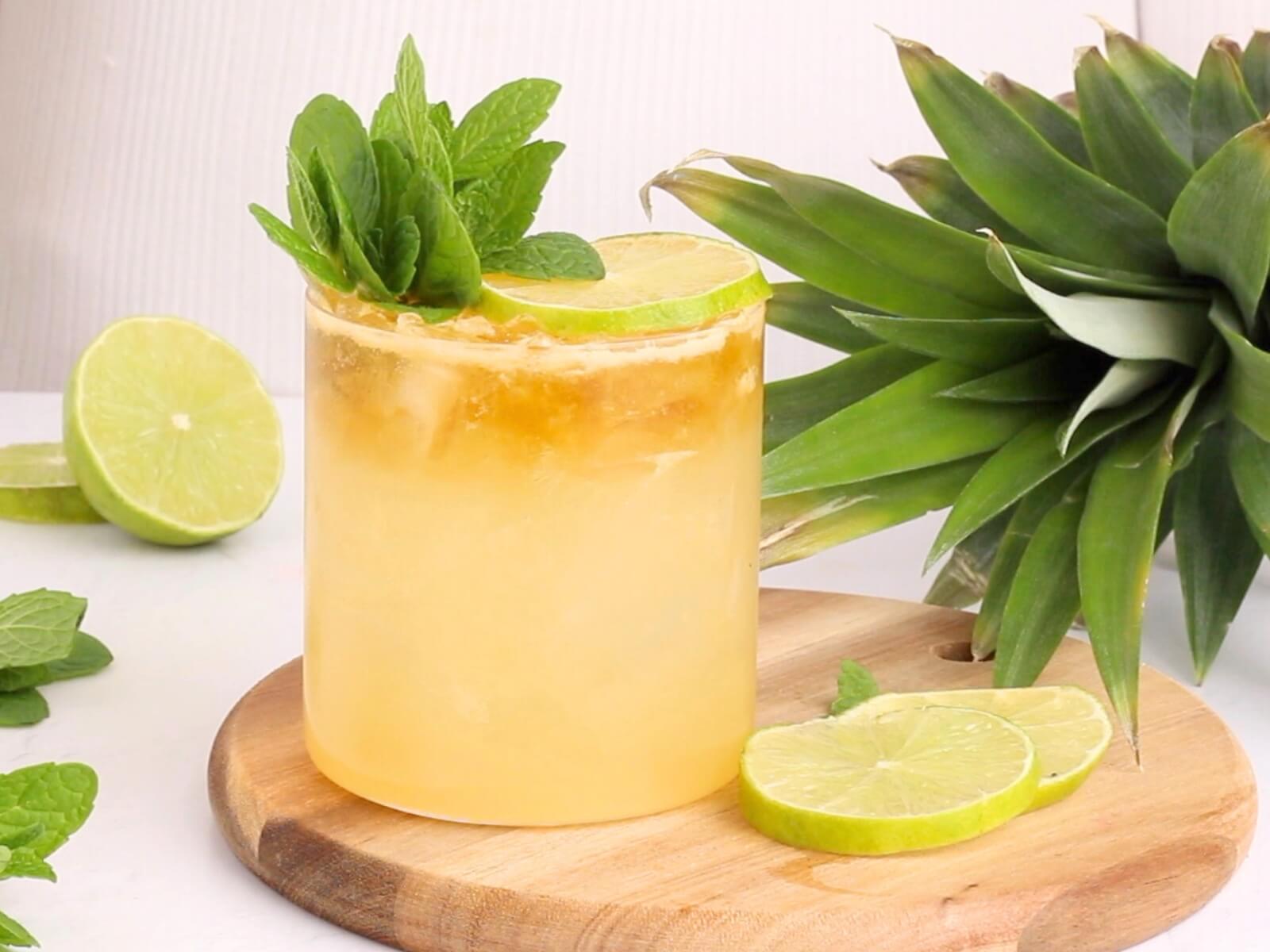 A refreshing yellow cocktail garnished with mint leaves and a lime slice, served in a glass on a wooden board next to lime slices and a fresh pineapple.