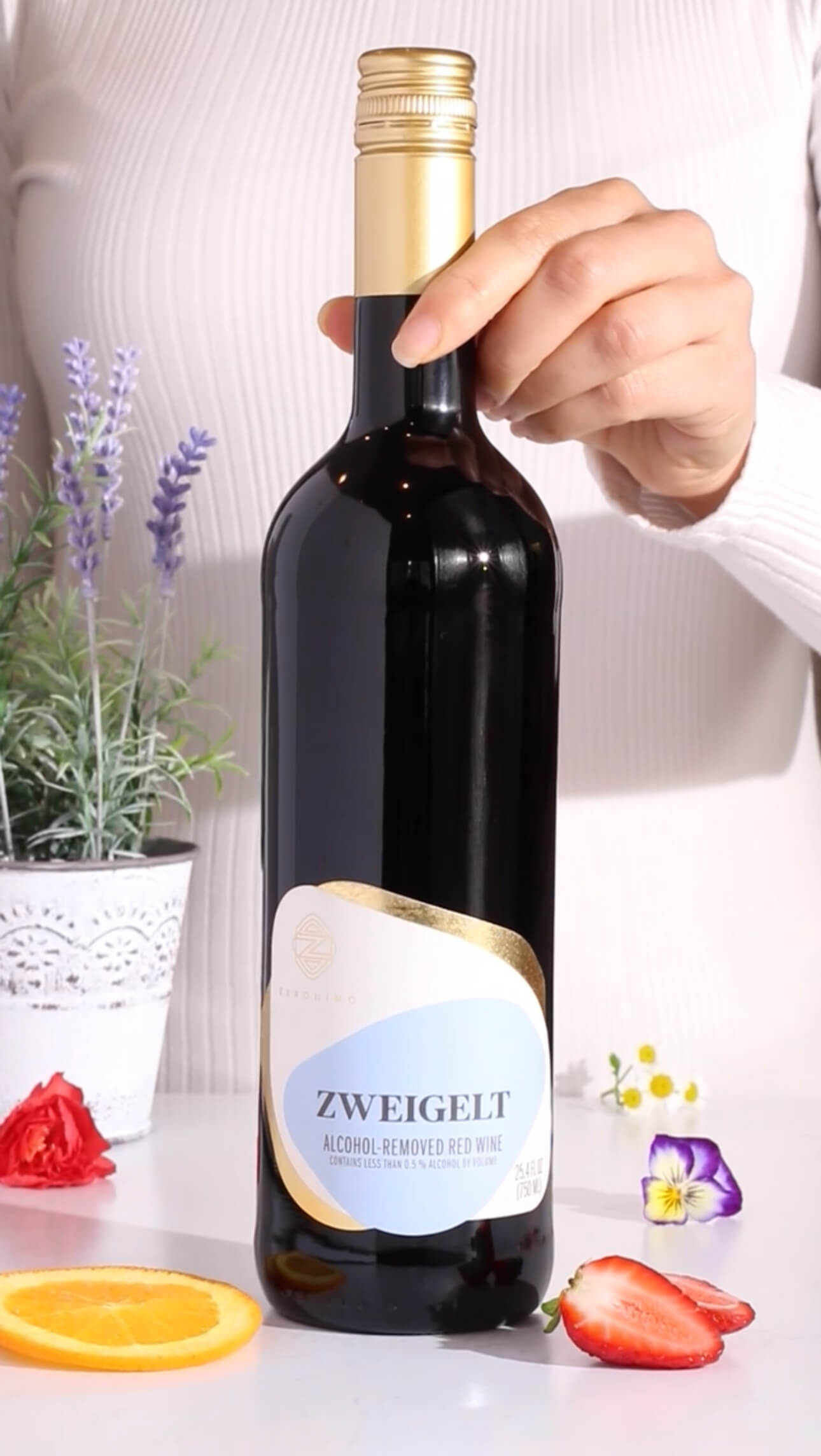 A hand holds a bottle of Zweigelt alcohol-removed red wine on a white table, with a potted lavender plant, flower petals, and orange slices nearby.
