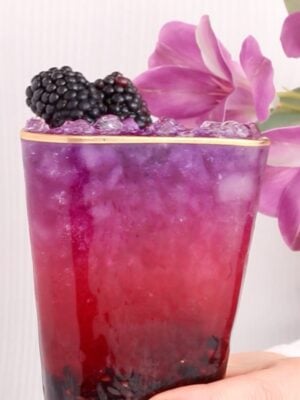 A hand holds a glass of vibrant, layered purple and pink halloween mocktail topped with ice and fresh blackberries, with pink flowers in the background.