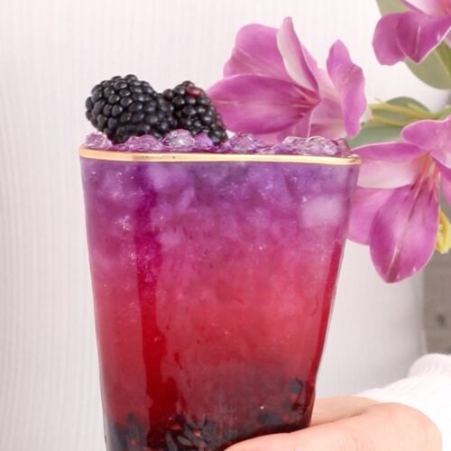 A hand holds a glass of vibrant, layered purple and pink halloween mocktail topped with ice and fresh blackberries, with pink flowers in the background.