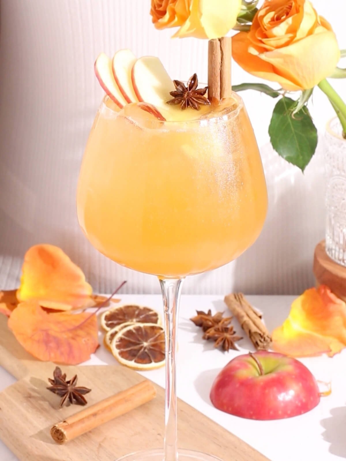 A large wine glass holds an orange cocktail garnished with apple slices, a cinnamon stick, and star anise. Orange roses, dried citrus, cinnamon sticks, star anise, and apple slices are arranged around the drink.