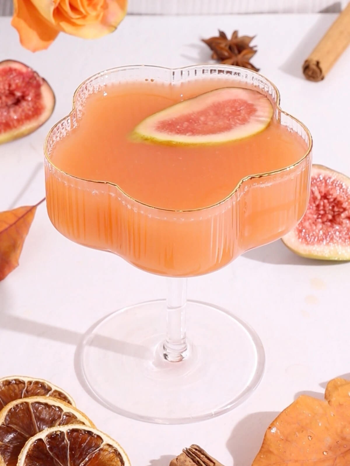 A pink fig mocktail garnished with a slice of fig is served in a flower-shaped glass. Dried citrus slices, fresh figs, a cinnamon stick, and autumn leaves are arranged around the glass on a white surface.