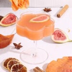 A peach-colored cocktail in a decorative glass, garnished with a fig slice. Surrounding the glass are figs, dried citrus, cinnamon sticks, star anise, an orange rose, and autumn leaves on a white surface.