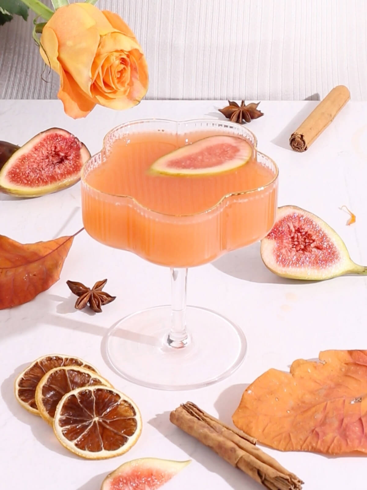 A peach-colored cocktail in a decorative glass, garnished with a fig slice. Surrounding the glass are figs, dried citrus, cinnamon sticks, star anise, an orange rose, and autumn leaves on a white surface.