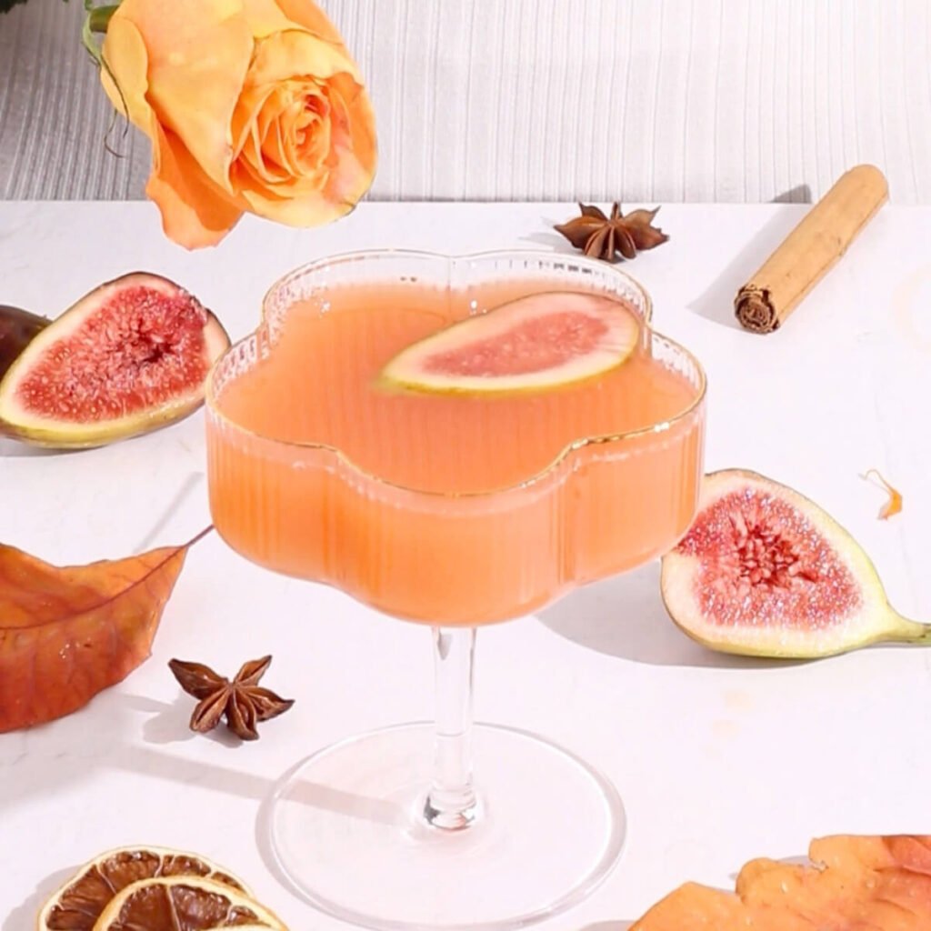 A pink-orange mocktail in a flower-shaped glass garnished with a fig slice, surrounded by figs, a cinnamon stick, star anise, dried citrus, an orange rose, and autumn leaves on a white surface.