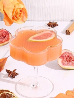 A pink-orange mocktail in a flower-shaped glass garnished with a fig slice, surrounded by figs, a cinnamon stick, star anise, dried citrus, an orange rose, and autumn leaves on a white surface.