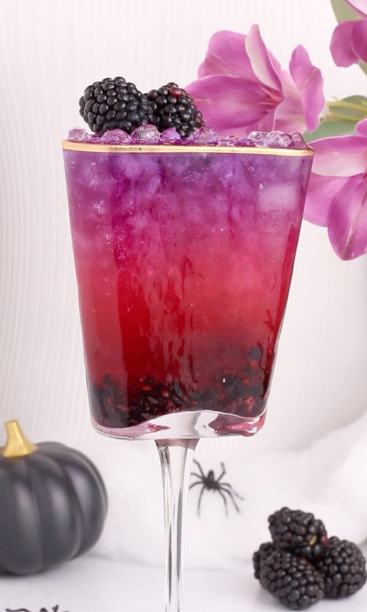A tall glass filled with a layered purple and red drink, topped with blackberries and crushed ice. There is a pink flower on the rim, a black pumpkin, a plastic spider, and blackberries in the background.