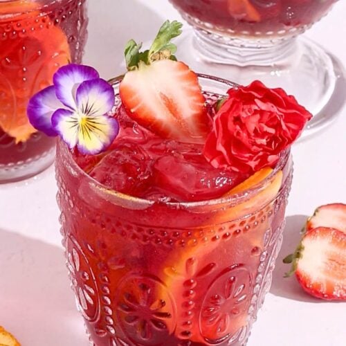 A glass of sparkling hibiscus sangria, garnished with a sliced strawberry, orange slices, a red carnation, and a purple and yellow pansy flower in an ornate glass. Another similar glass and sliced strawberries are in the background.