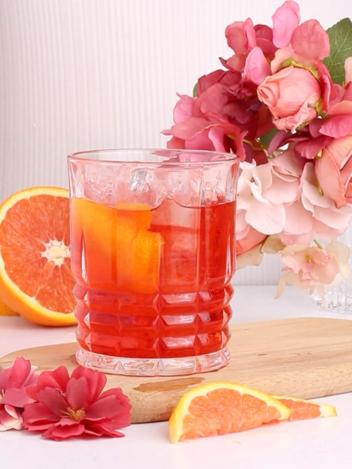 A glass of red cocktail with ice and an orange peel sits on a wooden board, surrounded by sliced oranges and pink flowers, against a light background.