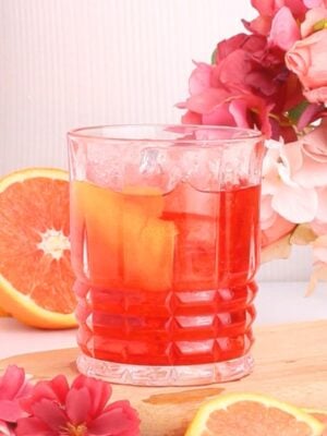 A non alcoholic negroni with ice and an orange slice garnish sits on a wooden board, surrounded by pink flowers and grapefruit slices.