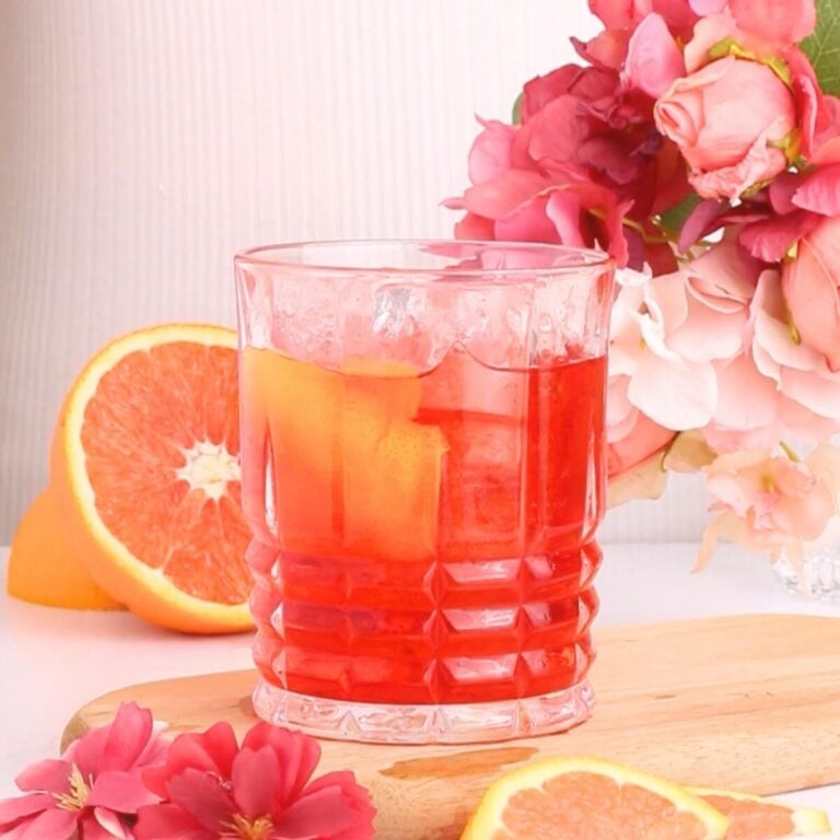 A non alcoholic negroni with ice and an orange slice garnish sits on a wooden board, surrounded by pink flowers and grapefruit slices.