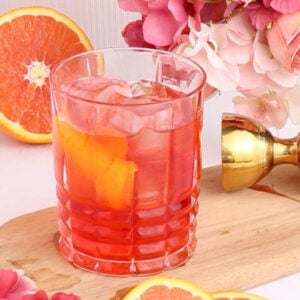A glass of pink mocktail with ice and orange slices sits on a wooden board, surrounded by pink flowers, orange slices, and a gold jigger in the background.