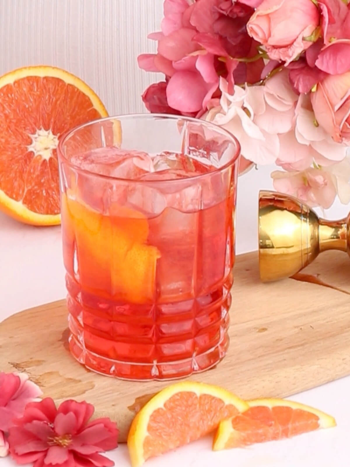 A glass of pink mocktail with ice and orange slices sits on a wooden board, surrounded by pink flowers, orange slices, and a gold jigger in the background.