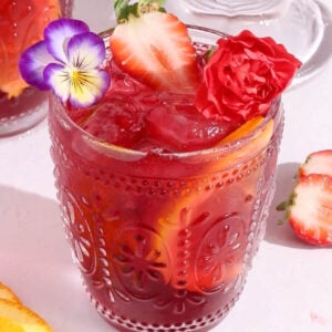 A decorative glass of sparkling hibiscus sangria with ice, orange and strawberry slices, topped with edible flowers and a red carnation, set on a white surface with scattered fruit and flowers around.