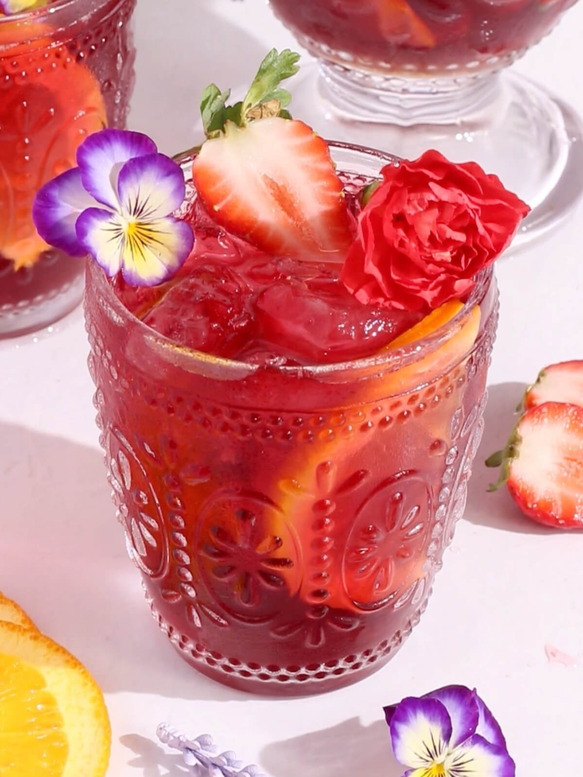 A decorative glass of sparkling hibiscus sangria with ice, orange and strawberry slices, topped with edible flowers and a red carnation, set on a white surface with scattered fruit and flowers around.