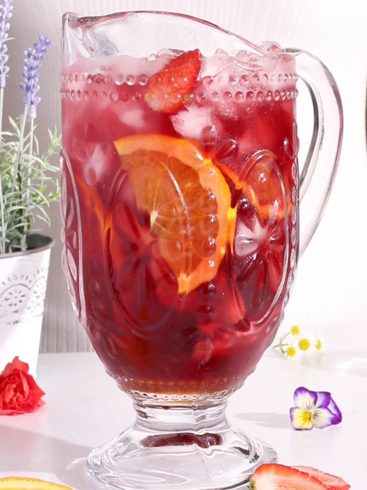 A glass pitcher filled with vibrant, sparkling hibiscus sangria, chilled with ice and garnished with orange and strawberry slices. The ornate floral design complements the fresh flowers and potted plant in the background on a white surface.