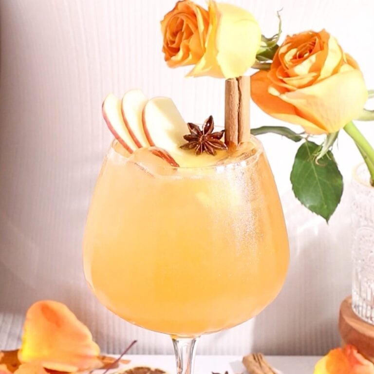A glass of orange-colored cocktail garnished with apple slices, a cinnamon stick, and star anise, sits in front of orange roses with flower petals scattered around.