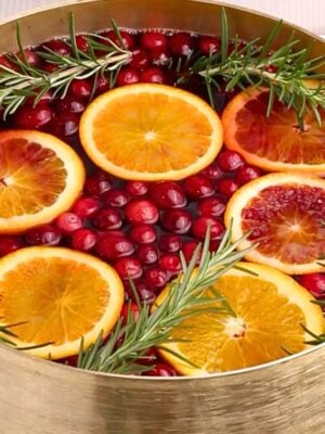 A gold punch bowl filled with water, whole cranberries, orange slices, and sprigs of fresh rosemary, creating a festive and colorful arrangement.