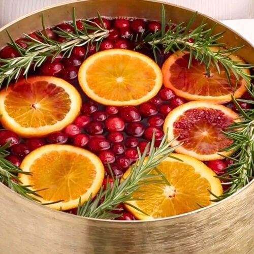 A gold punch bowl filled with water, whole cranberries, orange slices, and sprigs of fresh rosemary, creating a festive and colorful arrangement.