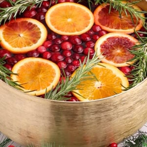 A gold punch bowl filled with cranberry juice, orange slices, and sprigs of rosemary, surrounded by festive greenery and red berries.