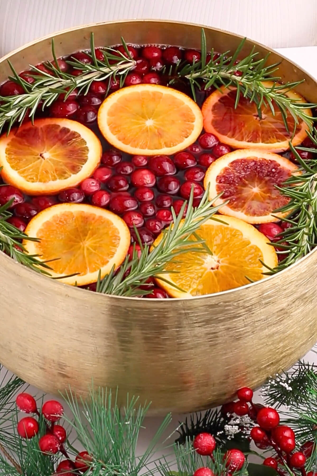 A gold punch bowl filled with cranberry juice, orange slices, and sprigs of rosemary, surrounded by festive greenery and red berries.