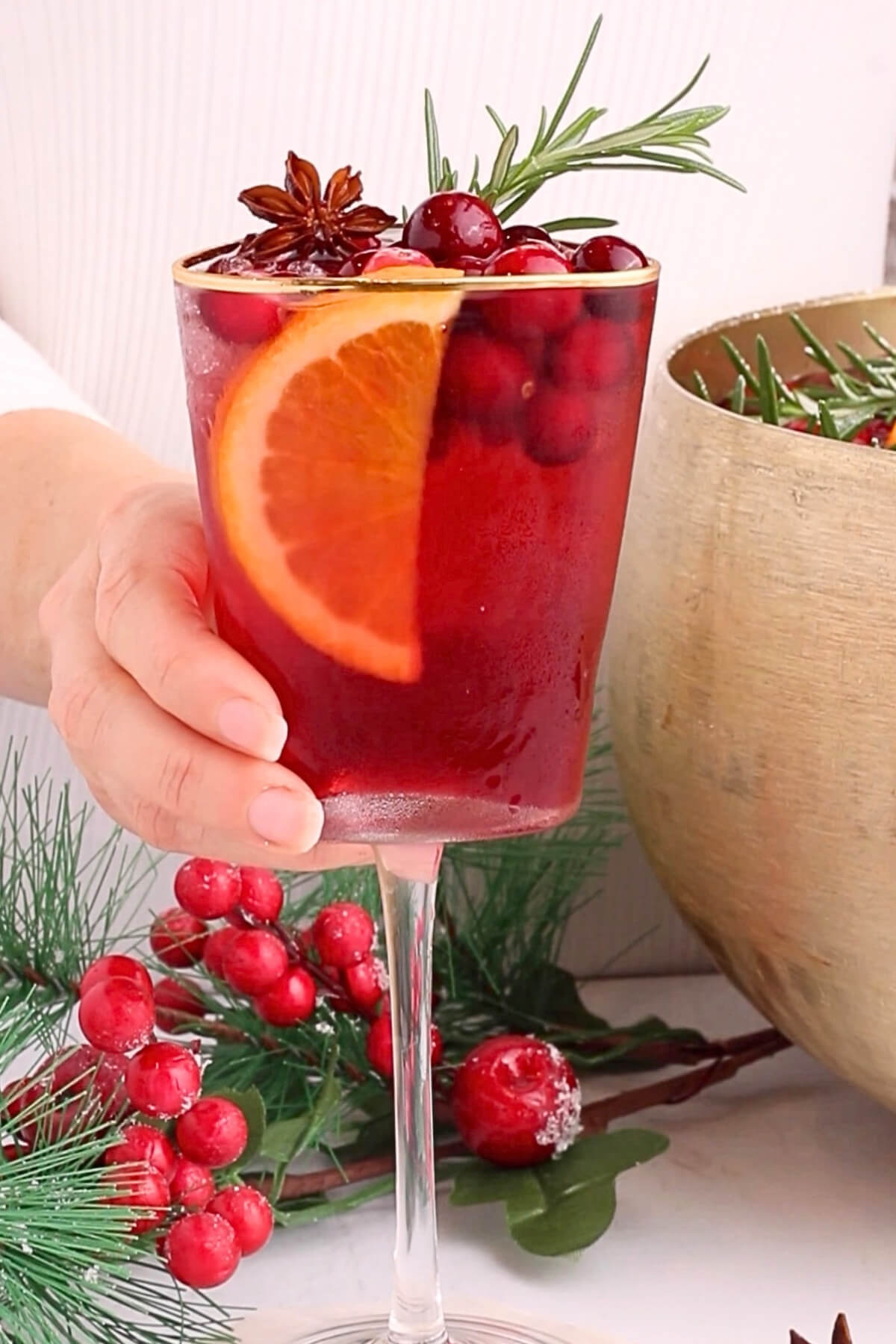 A hand holds a glass of red holiday punch garnished with an orange slice, cranberries, rosemary, and star anise, with festive greenery and red berries in the background.