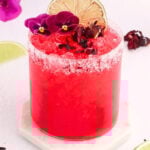 A vibrant red hibiscus margarita mocktail in a salt-rimmed glass is garnished with lime slices, purple flowers, and hibiscus petals, set on a white coaster with fresh lime wedges and flower petals nearby.