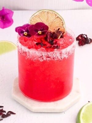 A vibrant red hibiscus margarita mocktail in a salt-rimmed glass is garnished with lime slices, purple flowers, and hibiscus petals, set on a white coaster with fresh lime wedges and flower petals nearby.