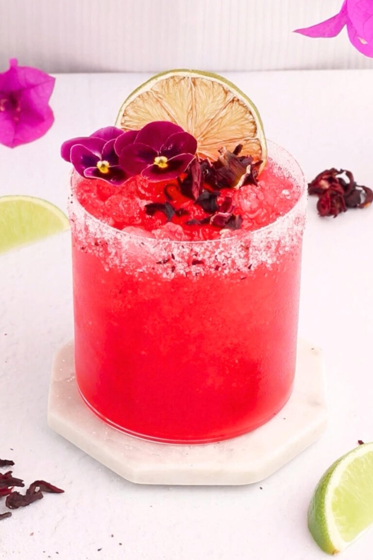 A vibrant red hibiscus margarita mocktail in a salt-rimmed glass is garnished with lime slices, purple flowers, and hibiscus petals, set on a white coaster with fresh lime wedges and flower petals nearby.