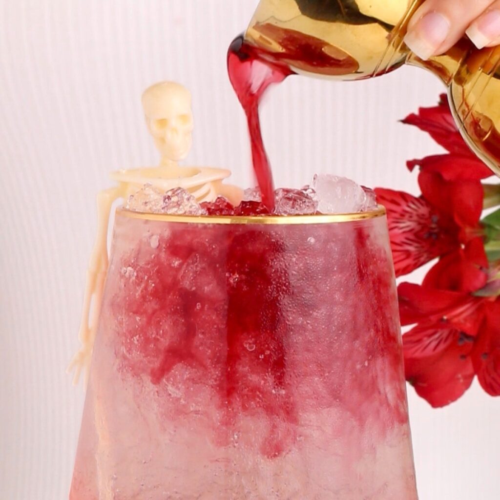 A gold pitcher pours red liquid over ice in a glass. A small plastic skeleton leans on the glass beside a red flower in the background.