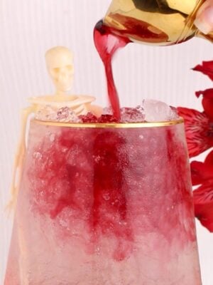 A gold pitcher pours red liquid over ice in a glass. A small plastic skeleton leans on the glass beside a red flower in the background.