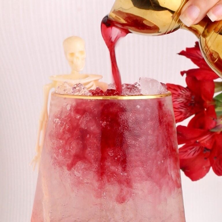 A gold pitcher pours red liquid over ice in a glass. A small plastic skeleton leans on the glass beside a red flower in the background.
