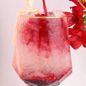 A glass filled with crushed ice and red liquid being poured in, with pomegranate seeds at the bottom. A small skeleton figurine decorates the rim, and red flowers are visible in the background.