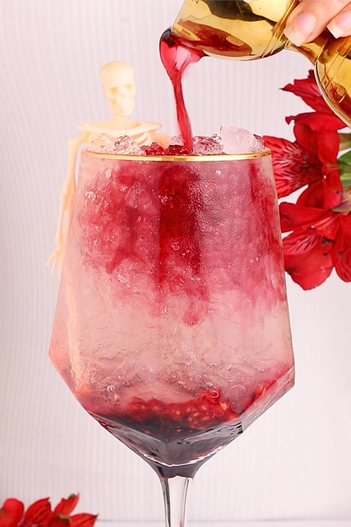 A glass filled with crushed ice and red liquid being poured in, with pomegranate seeds at the bottom. A small skeleton figurine decorates the rim, and red flowers are visible in the background.