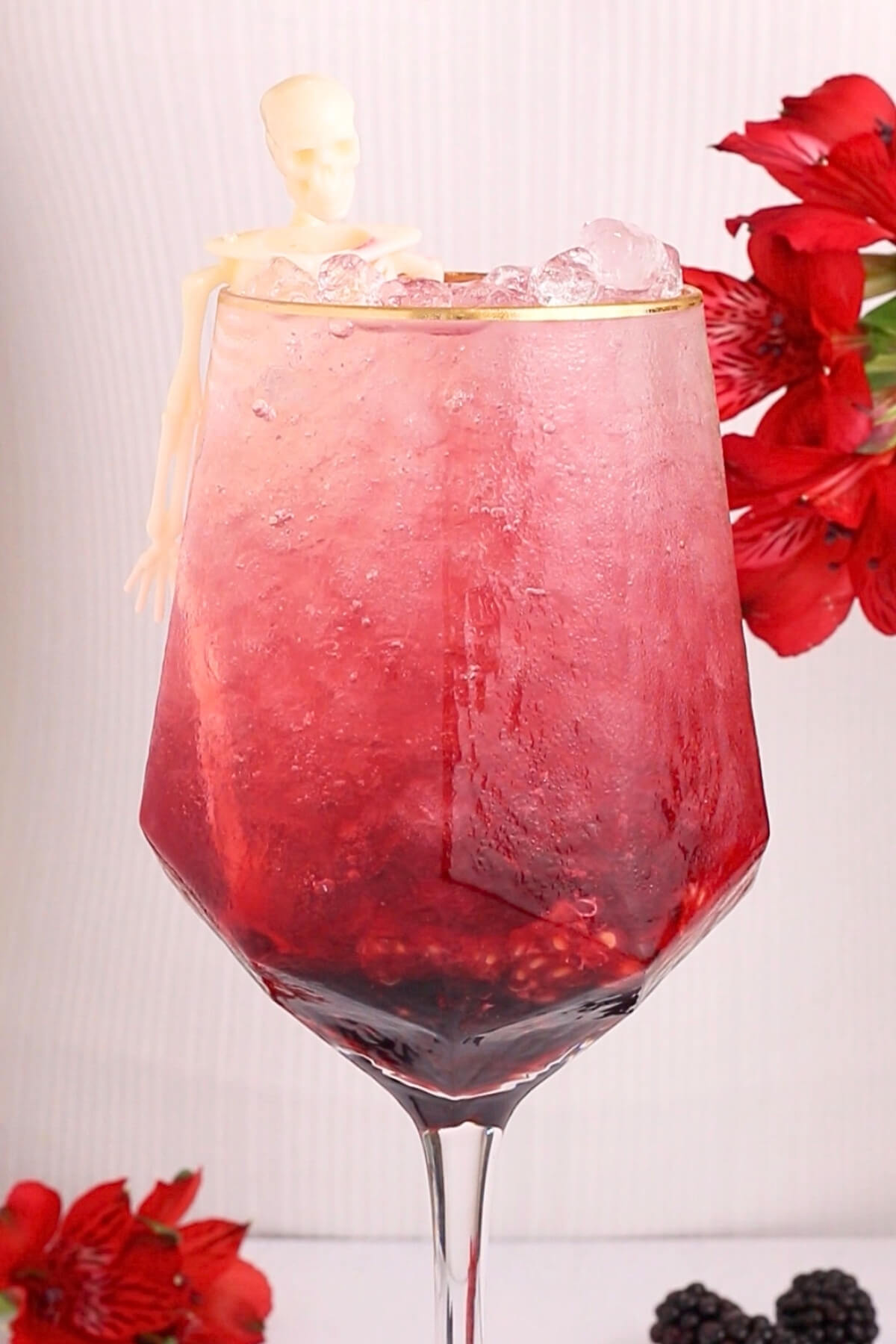 A large spooky mocktail with a skeleton in a wine glass with a red and pink icy drink. Red flowers and blackberries are placed in the background and foreground.