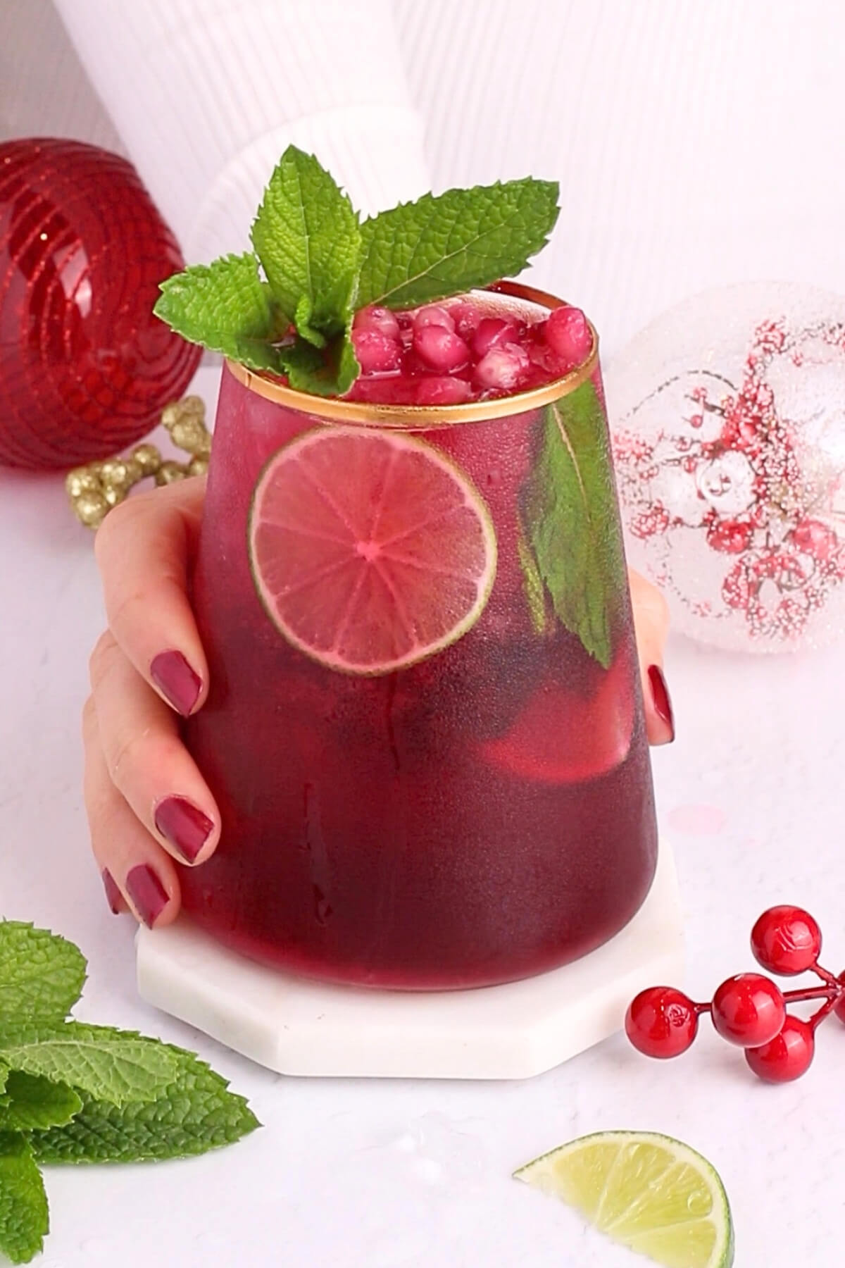 A hand with red-painted nails holds a christmas mojito mocktail garnished with mint leaves, cranberries, and lime slices. The background features festive holiday ornaments and scattered cranberries and mint leaves.