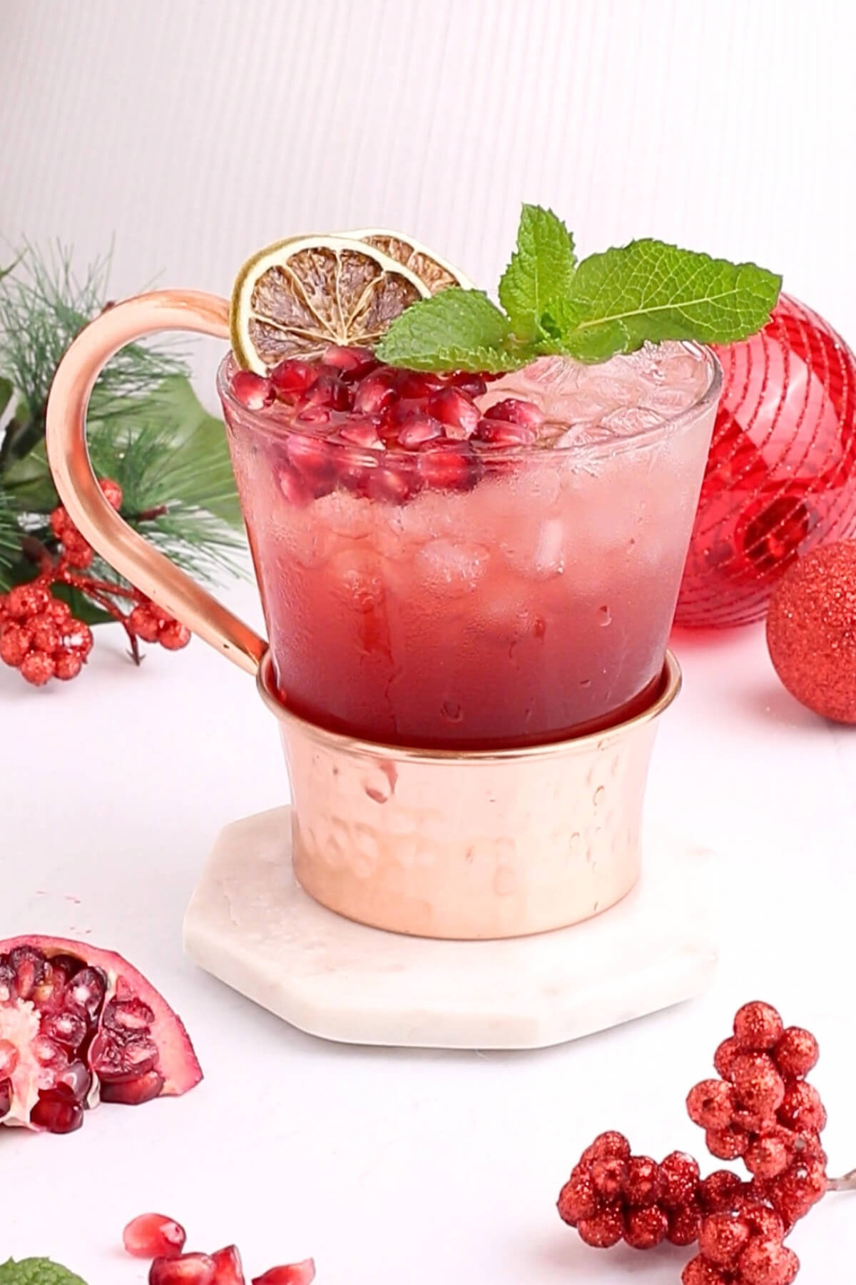 A festive drink in a copper mug topped with ice, pomegranate seeds, a dried lime slice, and a sprig of mint. The mug sits on a marble coaster with decorative berries and a pomegranate nearby.