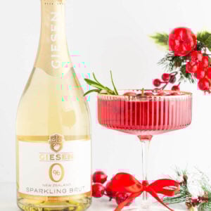 A bottle of Giesen sparkling brut stands beside a festive cocktail garnished with cranberries and rosemary, surrounded by dried oranges, red ribbon, gold berries, and holiday greenery on a white background.
