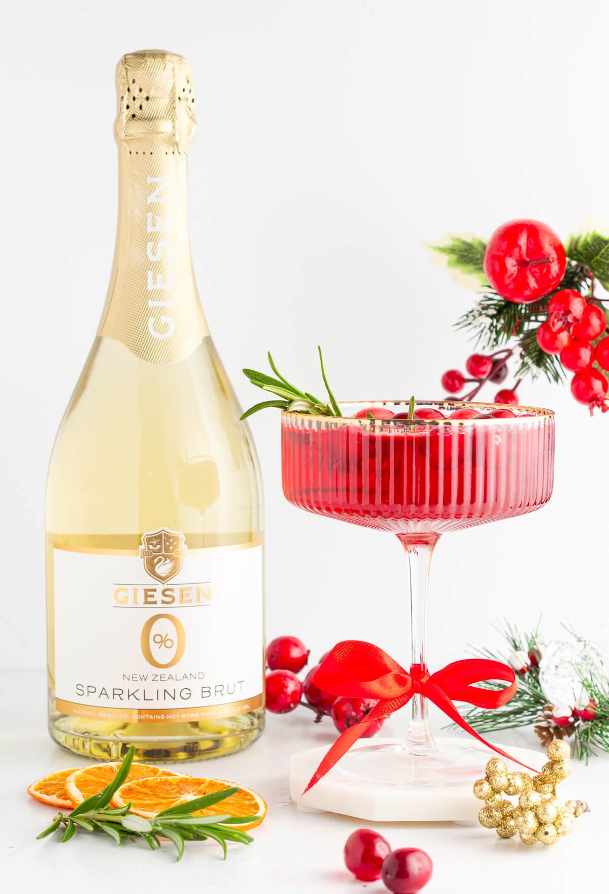 A bottle of Giesen sparkling brut stands beside a festive cocktail garnished with cranberries and rosemary, surrounded by dried oranges, red ribbon, gold berries, and holiday greenery on a white background.