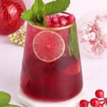 A vibrant red drink with lime slices, pomegranate seeds, and mint leaves in a glass. Red ornaments and gold beads are in the background, with mint sprigs and a lime wedge beside the glass.