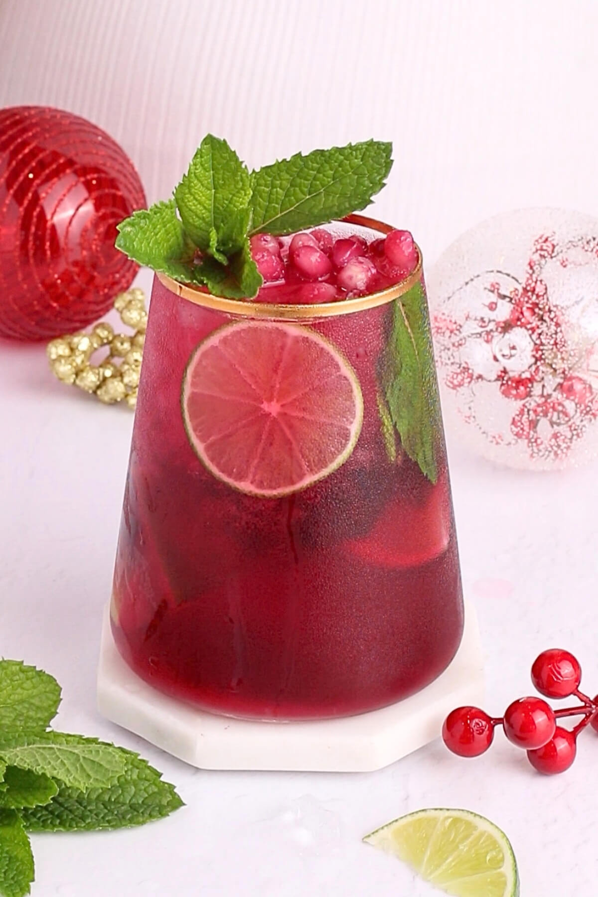 A vibrant red drink with lime slices, pomegranate seeds, and mint leaves in a glass. Red ornaments and gold beads are in the background, with mint sprigs and a lime wedge beside the glass.