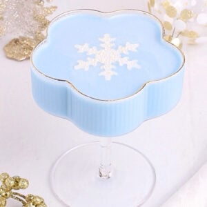 A blue Jack Frost mocktail in a flower-shaped glass, topped with a white glittery snowflake decoration. The drink is surrounded by gold and white festive holiday ornaments on a white surface.
