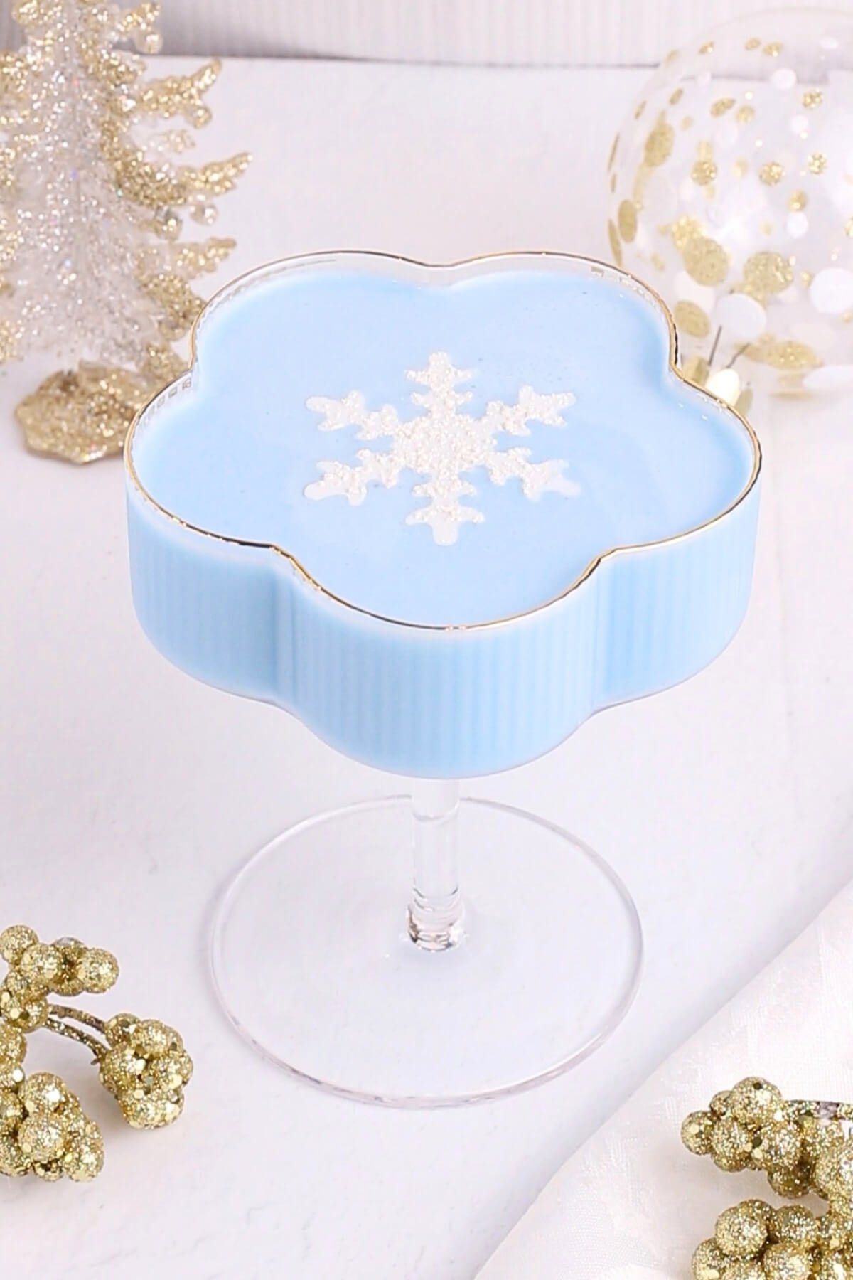 A blue Jack Frost mocktail in a flower-shaped glass, topped with a white glittery snowflake decoration. The drink is surrounded by gold and white festive holiday ornaments on a white surface.