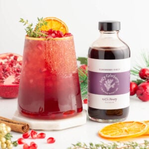 A glass of red punch garnished with orange slice, pomegranate seeds, and herbs sits beside a bottle of elderberry syrup. Pomegranate, cinnamon sticks, dried citrus, and festive decorations are in the background.